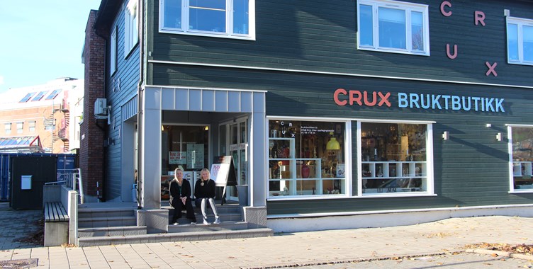 Grønt to etasjers hus med butikkvinduene til bruktbutikken og CRUX logo på huset. Det sitter to damer på trappen ved inngangen til bruktbutikken.lokalene